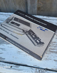 A boxed mini pneumatic pocket camera product package placed on a weathered white-painted wooden surface. The box shows product imagery and text including 'Mini Pneumatic Pocket Camera' and 'Take Me Along.