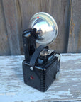 A vintage black wall-mounted spotlight with a polished silver dome reflector, photographed against a weathered wooden wall with peeling white paint.