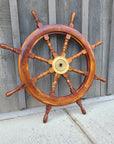 A wooden ship's wheel with a polished warm brown finish and a brass center hub, photographed against weathered gray wooden planks and a light concrete wall.