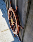 A decorative wooden ship's wheel wall hanging with a brass center medallion, shown leaning against a weathered wooden wall outdoors.