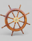 A wooden ship's wheel with a polished warm brown finish and a brass center hub, photographed against weathered gray wooden planks and a light concrete wall.