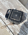 An antique black Rolle Rux camera with a textured leather front cover and silver metal accents, resting on a weathered, peeling white-painted wooden surface.