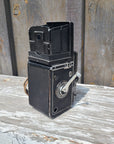 A vintage black metal camera body with a white hand crank handle, photographed mounted against weathered wooden boards.