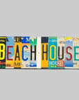 A custom license plate sign made from repurposed plates, mounted on painted wood with a variety of colorful text and graphics, spelling out 'BEACH HOUSE'.