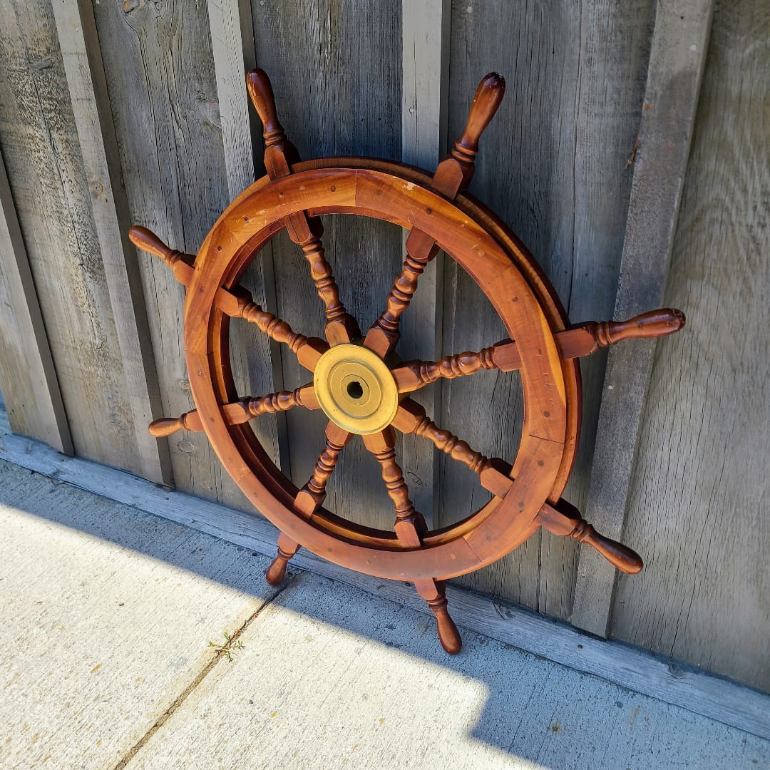 A large wooden ship's wheel with eight spokes and a brass center hub, leaning against a weathered gray wooden fence on a concrete surface.