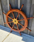 A large wooden ship's wheel with eight spokes and a brass center hub, leaning against a weathered gray wooden fence on a concrete surface.