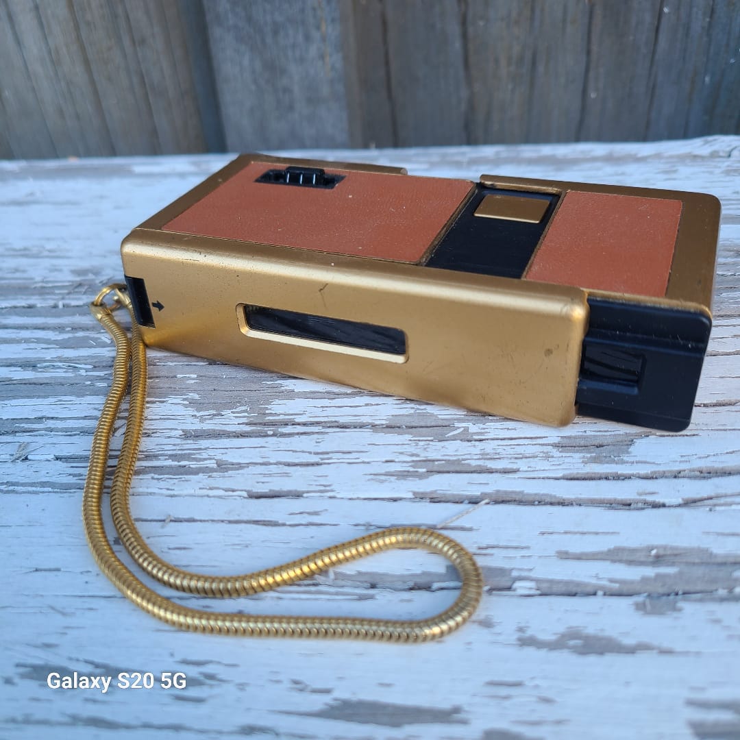 A vintage gold and terracotta orange instant camera with a coiled gold neck strap, resting on a weathered white wooden surface.