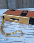 A vintage gold and terracotta orange instant camera with a coiled gold neck strap, resting on a weathered white wooden surface.