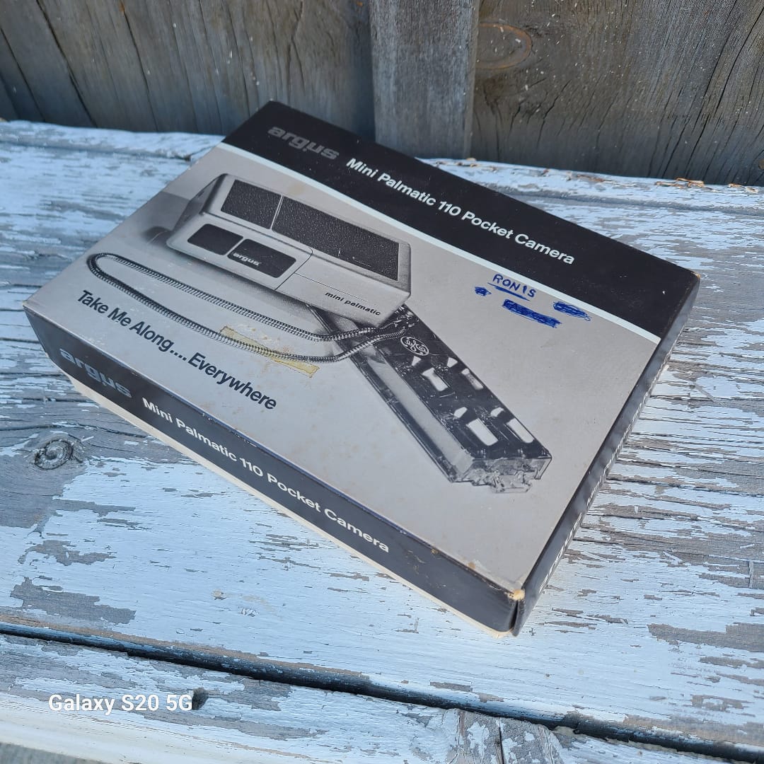 A boxed mini pneumatic pocket camera product package placed on a weathered white-painted wooden surface. The box shows product imagery and text including 'Mini Pneumatic Pocket Camera' and 'Take Me Along.