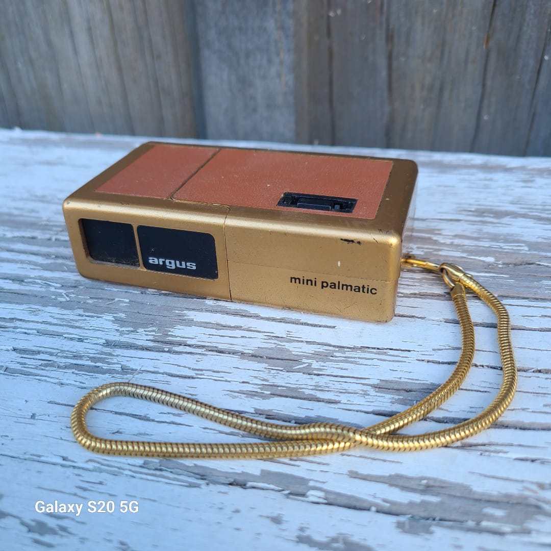 A mini argus palm-sized digital camera in gold and orange-brown, with a gold coiled wrist strap, resting on a weathered white-painted wooden surface.