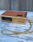 A mini argus palm-sized digital camera in gold and orange-brown, with a gold coiled wrist strap, resting on a weathered white-painted wooden surface.