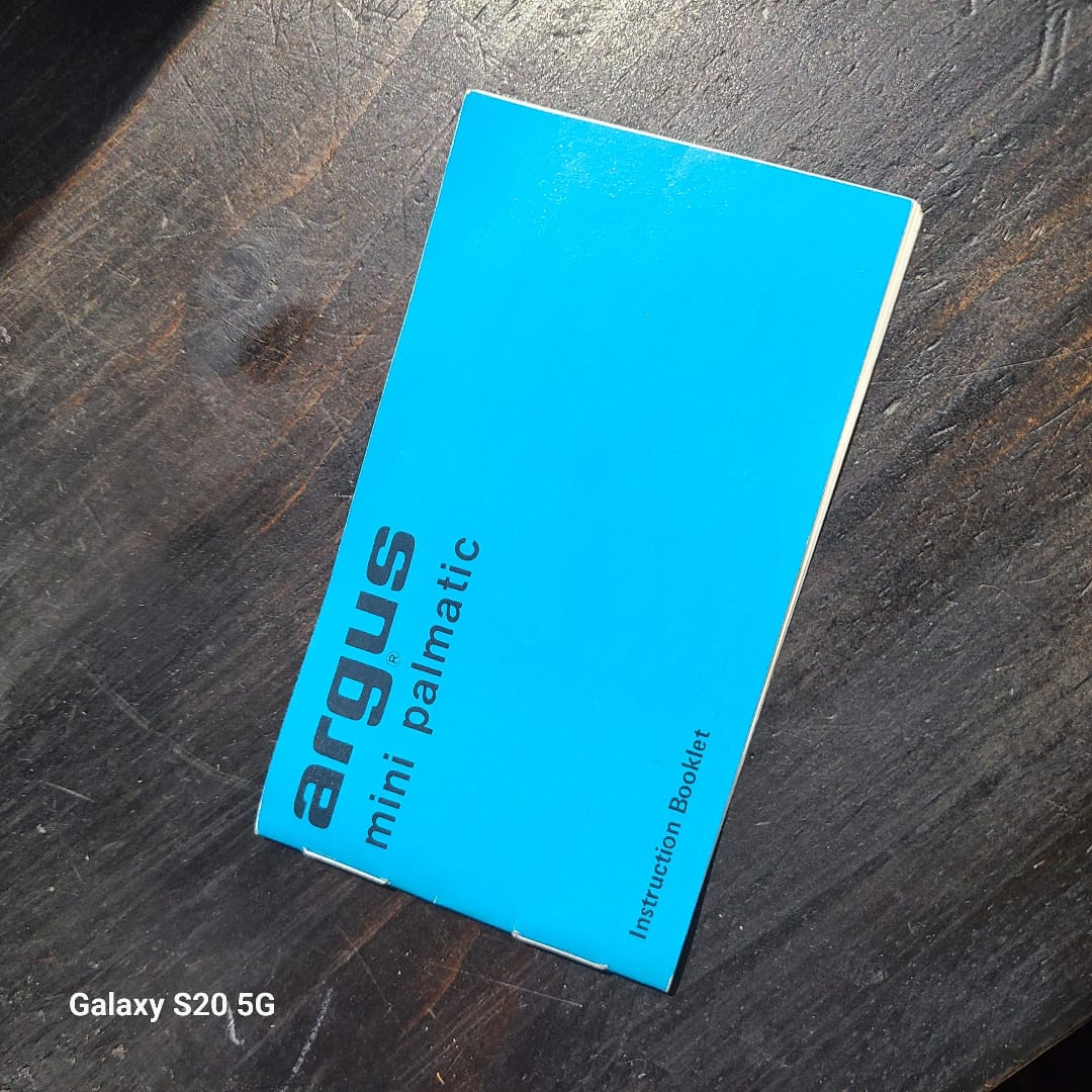 A bright blue Argus mini palmatic instruction booklet lies diagonally on a dark wooden surface. The text 'argus mini palmatic' is printed on the cover in black, and a small text watermark 'Galaxy S20 5G' is visible in the bottom-left corner.