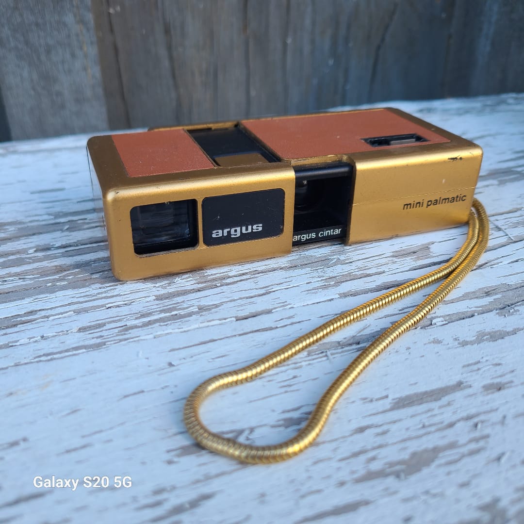 A vintage Argus mini instant camera in gold and orange-brown plastic with a gold wrist strap, resting on a weathered white wooden surface.