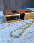 A vintage Argus mini instant camera in gold and orange-brown plastic with a gold wrist strap, resting on a weathered white wooden surface.