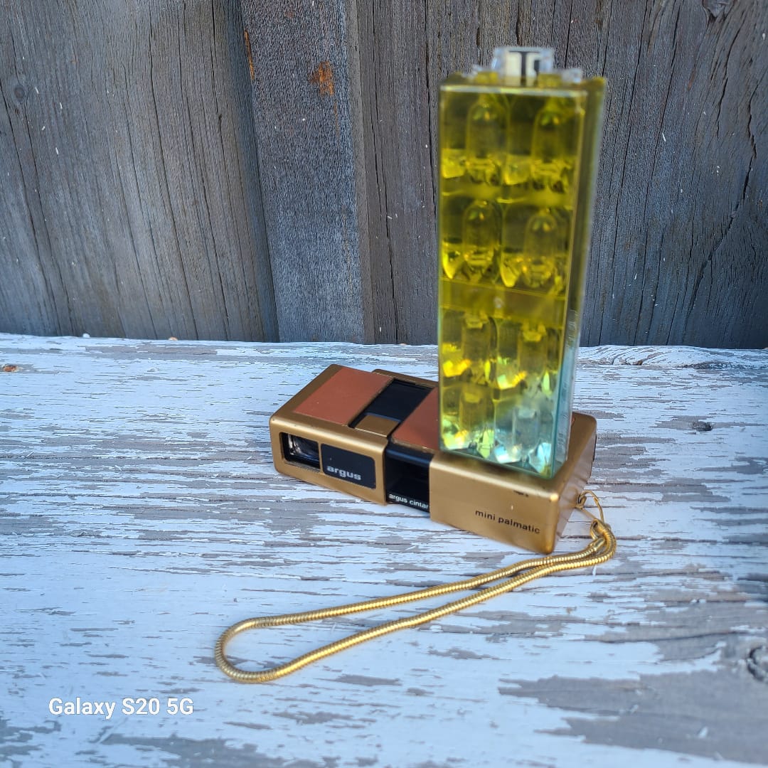 A novelty lighter shaped like a camera with a gold-tone body, black details, and a translucent yellow-green lens cover, attached to a gold wrist strap. It is photographed outdoors on a weathered white-painted wooden surface next to a gray wood plank background.