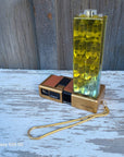 A novelty lighter shaped like a camera with a gold-tone body, black details, and a translucent yellow-green lens cover, attached to a gold wrist strap. It is photographed outdoors on a weathered white-painted wooden surface next to a gray wood plank background.