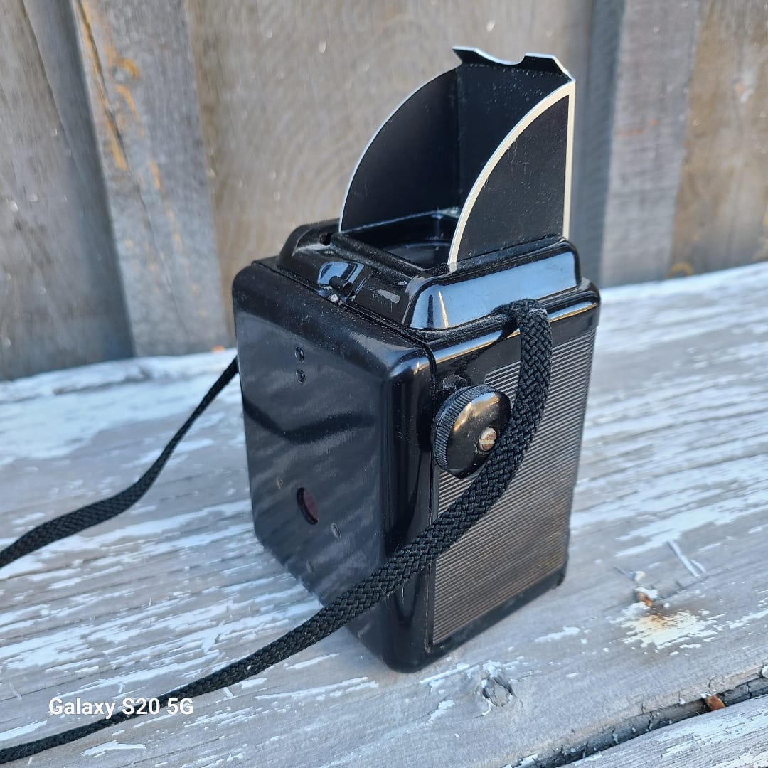 A vintage black camera with a textured top panel and a black woven strap, photographed on weathered wooden planks outdoors.