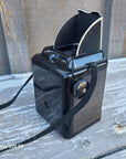 A vintage black camera with a textured top panel and a black woven strap, photographed on weathered wooden planks outdoors.