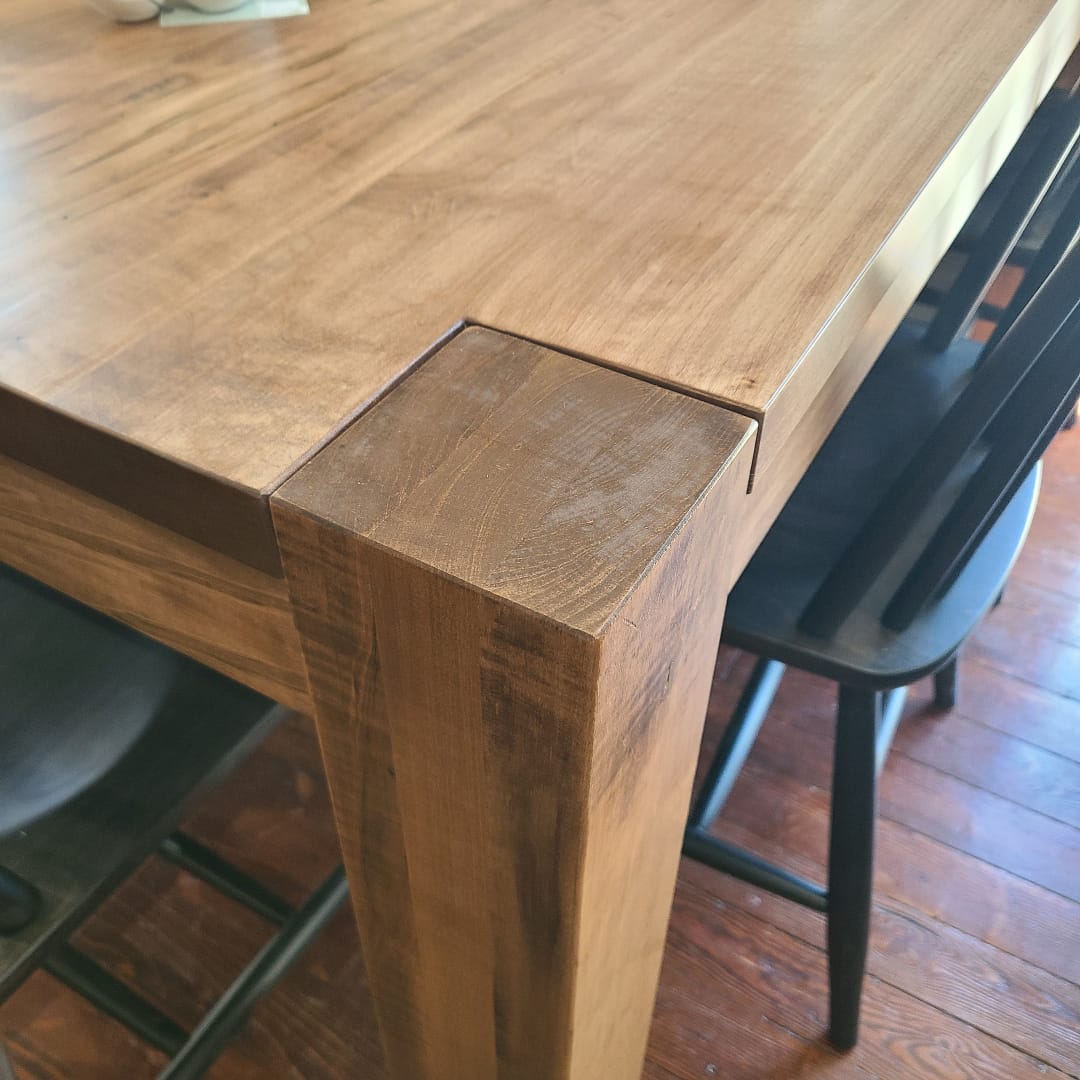 Close-up of a wooden dining table corner with a thick block joint and visible wood grain, shown next to black chairs on a hardwood floor.