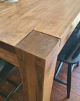 Close-up of a wooden dining table corner with a thick block joint and visible wood grain, shown next to black chairs on a hardwood floor.