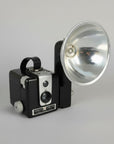 A vintage black and silver folding camera with an attached silver reflector, photographed on a neutral light gray background.