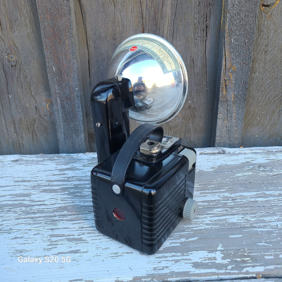 A vintage black wall-mounted spotlight with a polished silver dome reflector, photographed against a weathered wooden wall with peeling white paint.