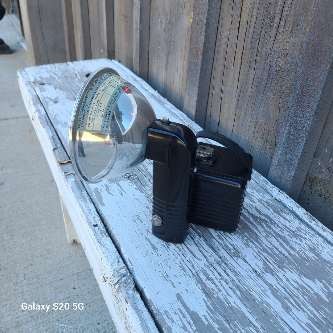 A black handheld spotlight with a clear reflective dome lens, photographed resting on a weathered white wooden surface outdoors.