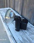 A black handheld spotlight with a clear reflective dome lens, photographed resting on a weathered white wooden surface outdoors.