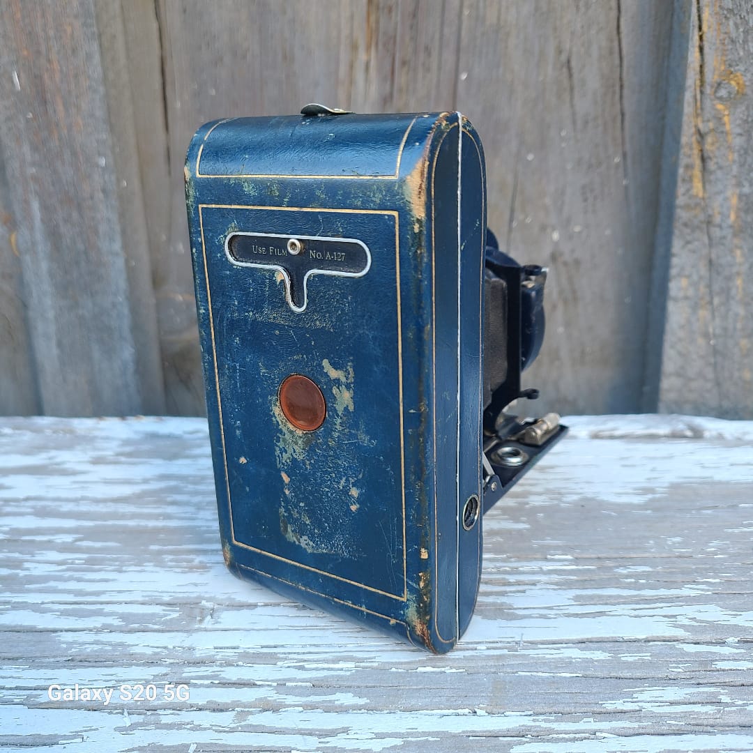 An antique blue leather camera case with gold trim, mounted on a weathered wooden wall next to a white-painted wood panel.