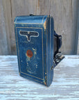 An antique blue leather camera case with gold trim, mounted on a weathered wooden wall next to a white-painted wood panel.