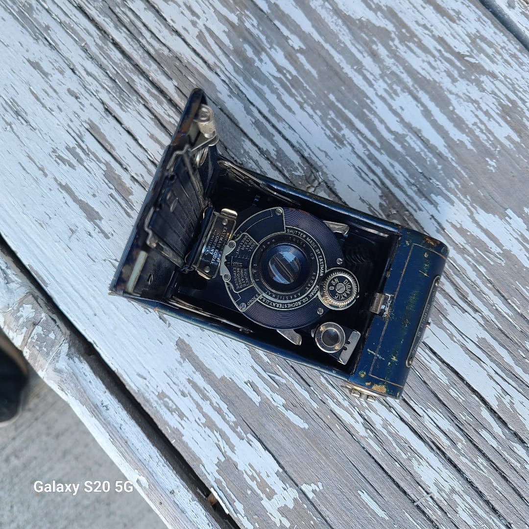 A vintage black and blue folding camera with silver metal details, shown open on a weathered, white-painted wooden surface.