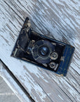 A vintage black and blue folding camera with silver metal details, shown open on a weathered, white-painted wooden surface.