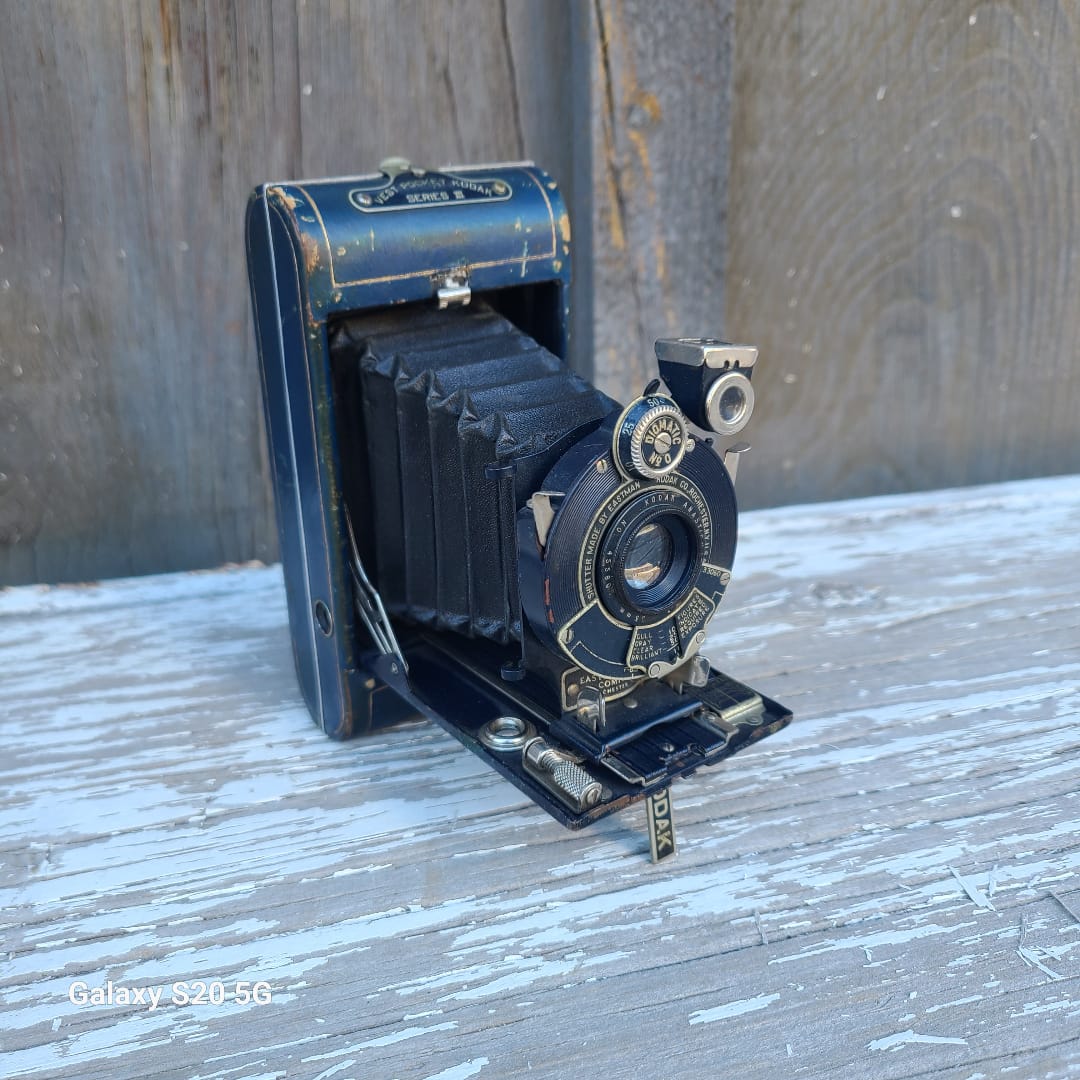 A vintage black and blue folding camera with silver metal dials, resting on a weathered wooden surface with peeling white paint.