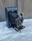 A vintage black and blue folding camera with silver metal dials, resting on a weathered wooden surface with peeling white paint.