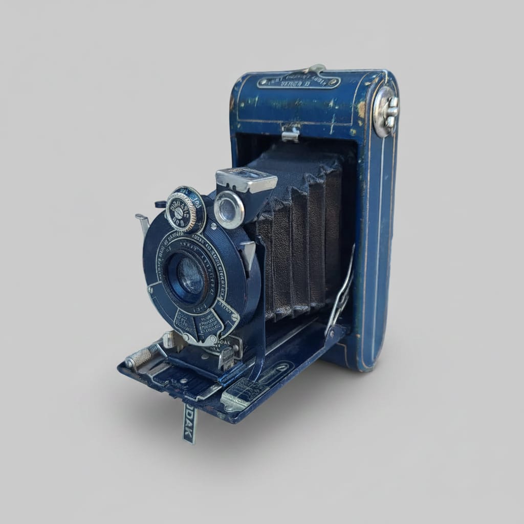 An antique blue folding camera with a black bellows, photographed resting against a weathered white-painted wooden surface.