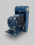 An antique blue folding camera with a black bellows, photographed resting against a weathered white-painted wooden surface.