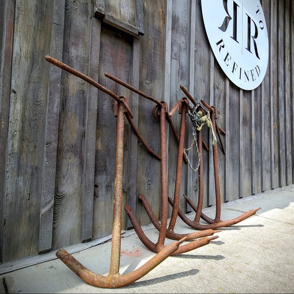 Rusty metal anchor-like props leaning against a weathered wooden building wall, with a round white sign reading 'Rustic To Refined' and a storefront entrance in the background.