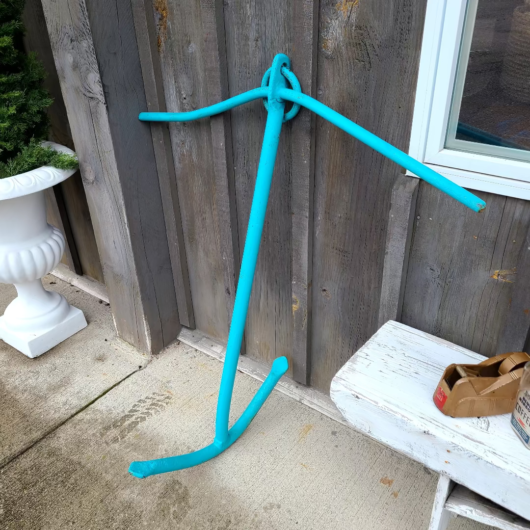 A bright turquoise blue anchor-shaped object leaning against a weathered gray wooden fence on a concrete patio, with a white bench and a planter visible nearby.