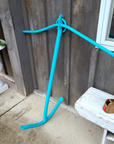 A bright turquoise blue anchor-shaped object leaning against a weathered gray wooden fence on a concrete patio, with a white bench and a planter visible nearby.