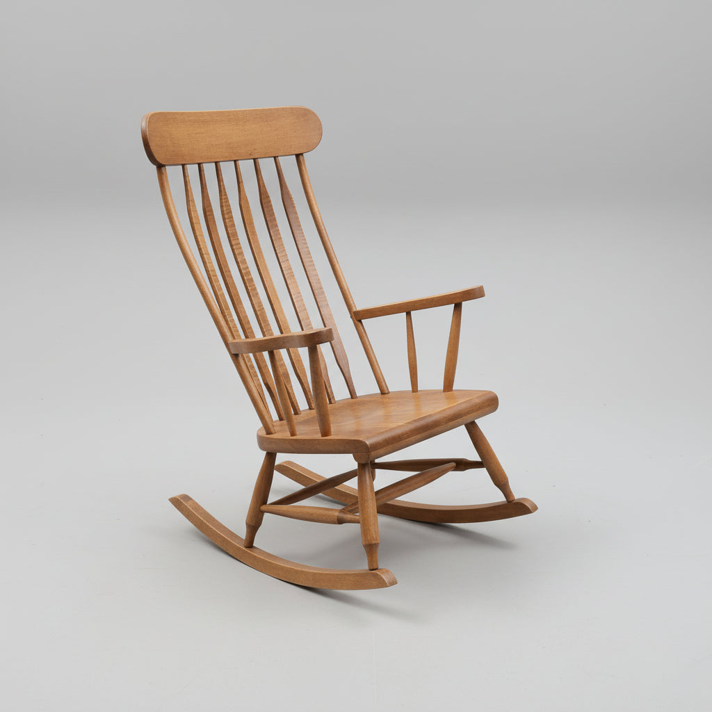 A traditional wooden rocking chair with a smooth finish and visible wood grain patterns. The chair has a scooped seat and a high backrest, and is designed with vertical slats between the seat and backrest.