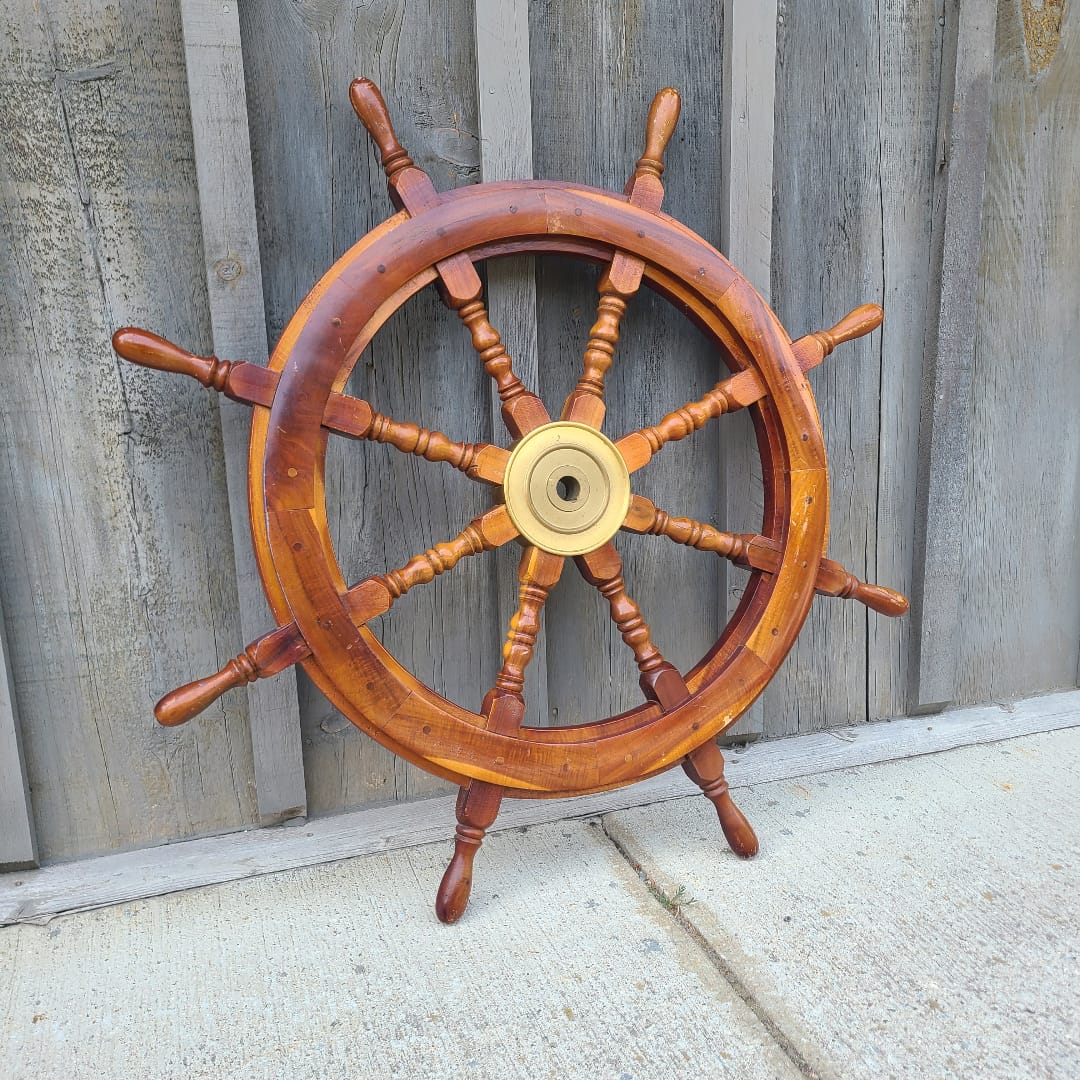 A wooden ship's wheel with a polished warm brown finish and a brass center hub, photographed against weathered gray wooden planks and a light concrete wall.