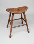 A wooden saddle stool with a smooth finish and turned leg, featuring a grab hole in the seat for easy mobility.