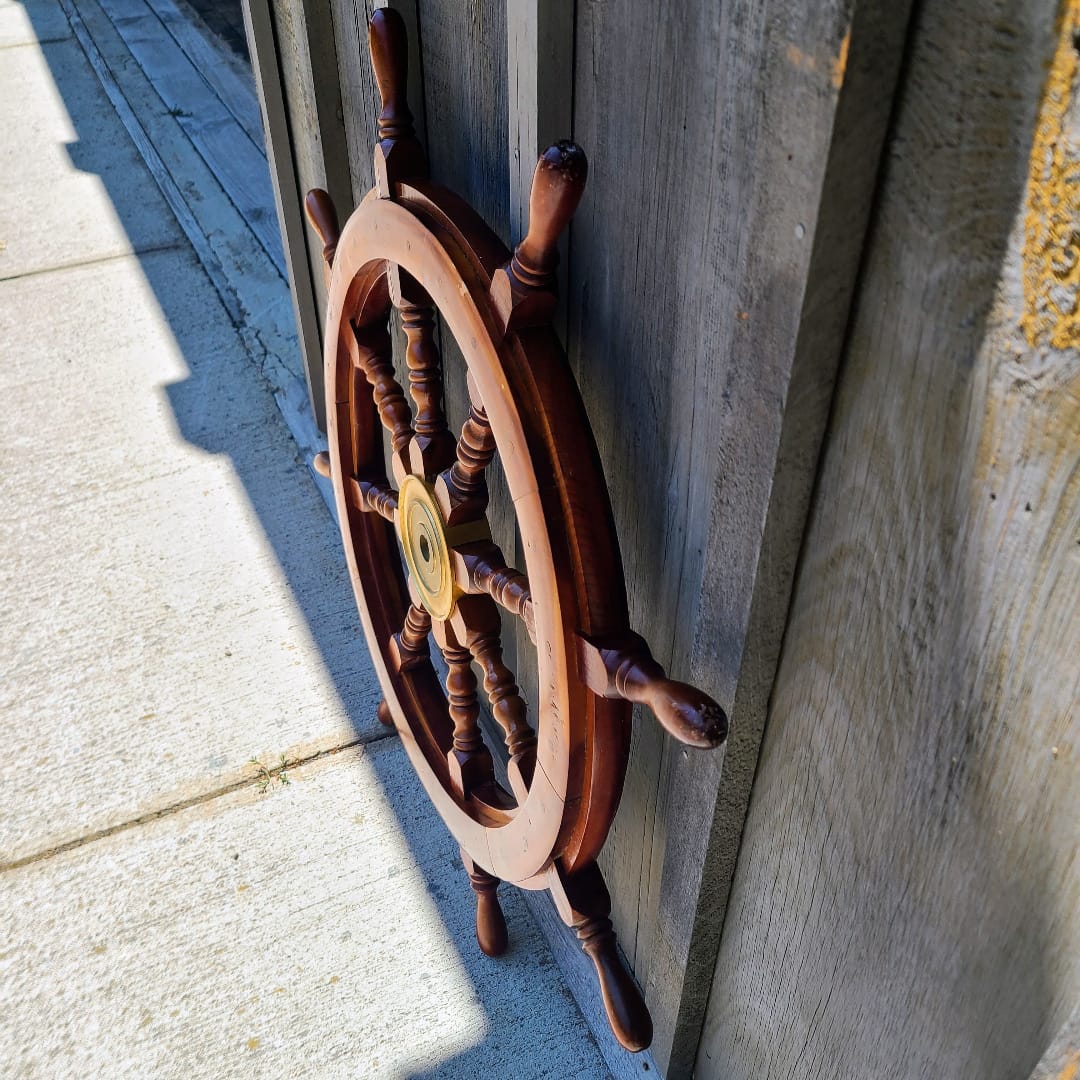 A decorative wooden ship's wheel wall hanging with a brass center medallion, shown leaning against a weathered wooden wall outdoors.