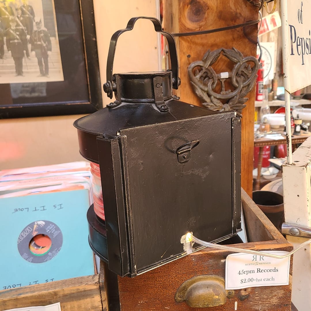 A vintage black metal lantern-style lamp with a handle and front panel, displayed on a wooden shelf in a shop setting with records and framed pictures in the background.