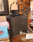A vintage black metal lantern-style lamp with a handle and front panel, displayed on a wooden shelf in a shop setting with records and framed pictures in the background.