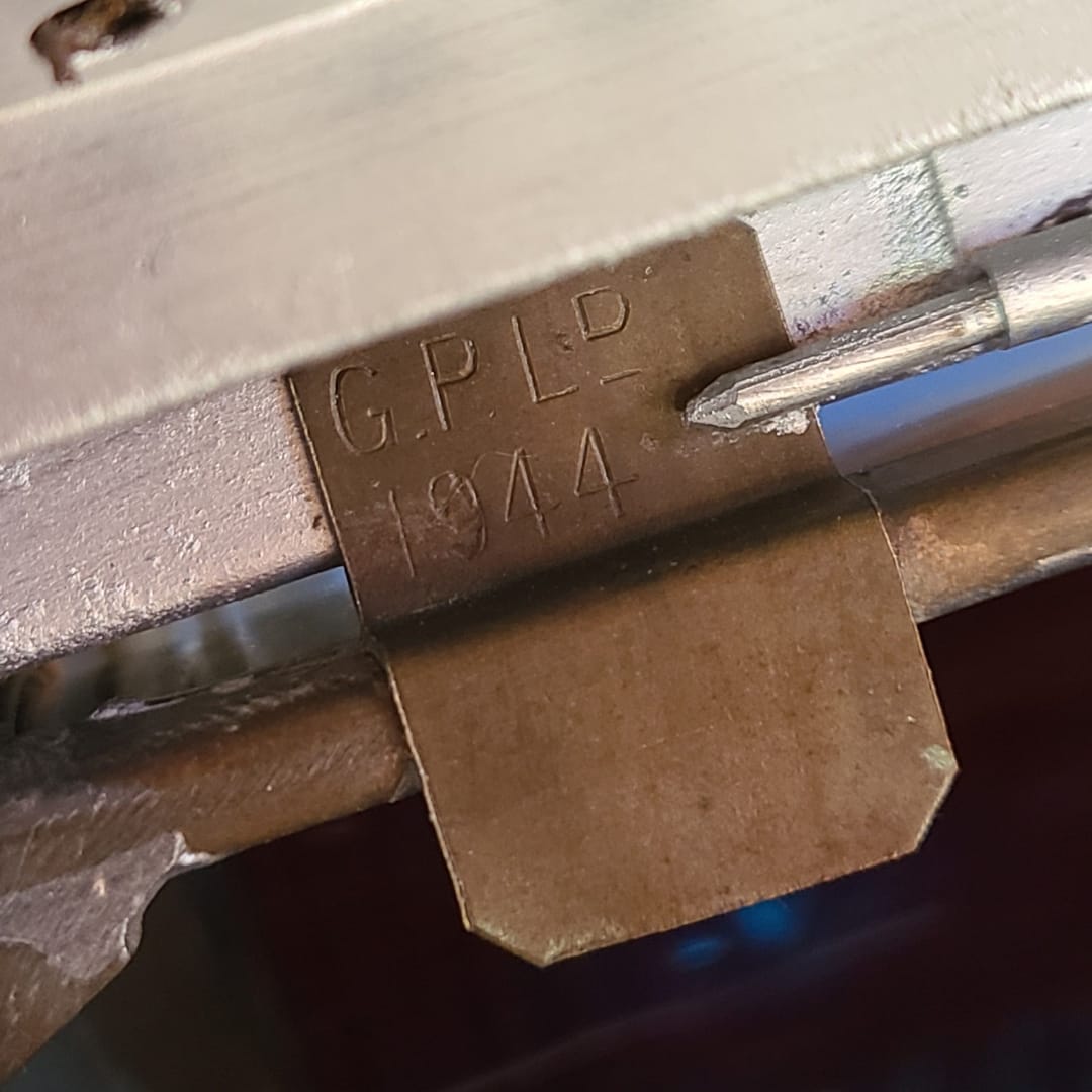 Close-up of a metal latch or lock mechanism with an embossed 'GPLD 1944' mark on a brass-colored tab, with a small metal tool touching the surface.