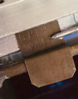 Close-up of a metal latch or lock mechanism with an embossed 'GPLD 1944' mark on a brass-colored tab, with a small metal tool touching the surface.