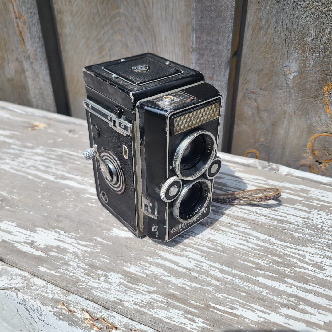 A vintage black double-lens reflex camera with silver metal dials and trim, photographed at an angle against a weathered white-painted wooden surface.