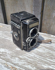 A vintage black double-lens reflex camera with silver metal dials and trim, photographed at an angle against a weathered white-painted wooden surface.