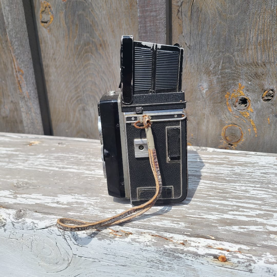 A vintage black and silver folding camera with a textured grip and a worn leather strap, photographed against a weathered wooden background.
